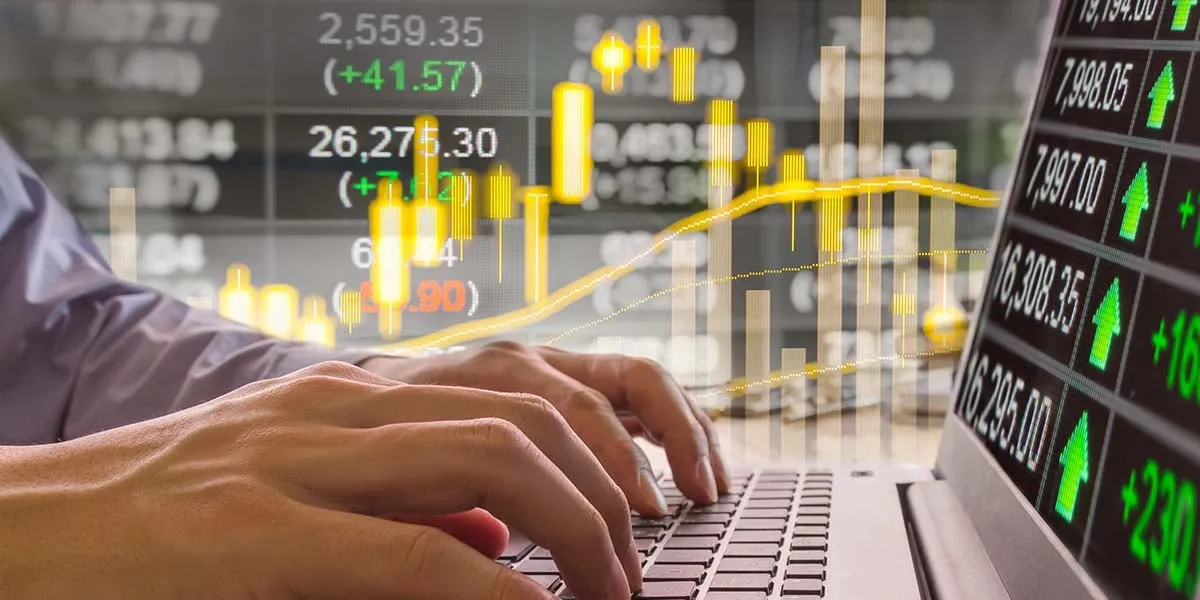 Hands typing on a laptop in front of a quote board and a price chart
